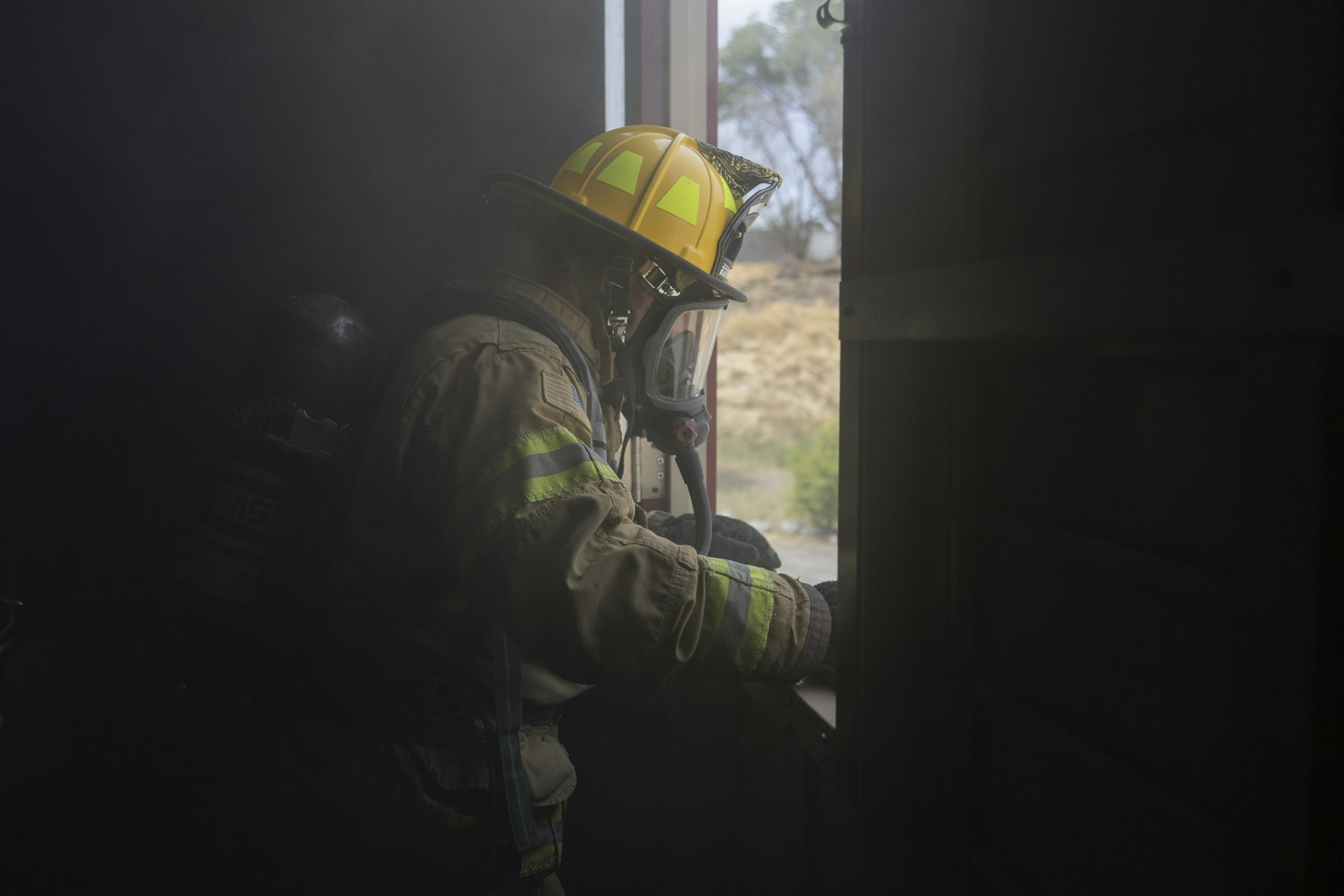 Firefighter in full gear looks out a window, surrounded by smoke.