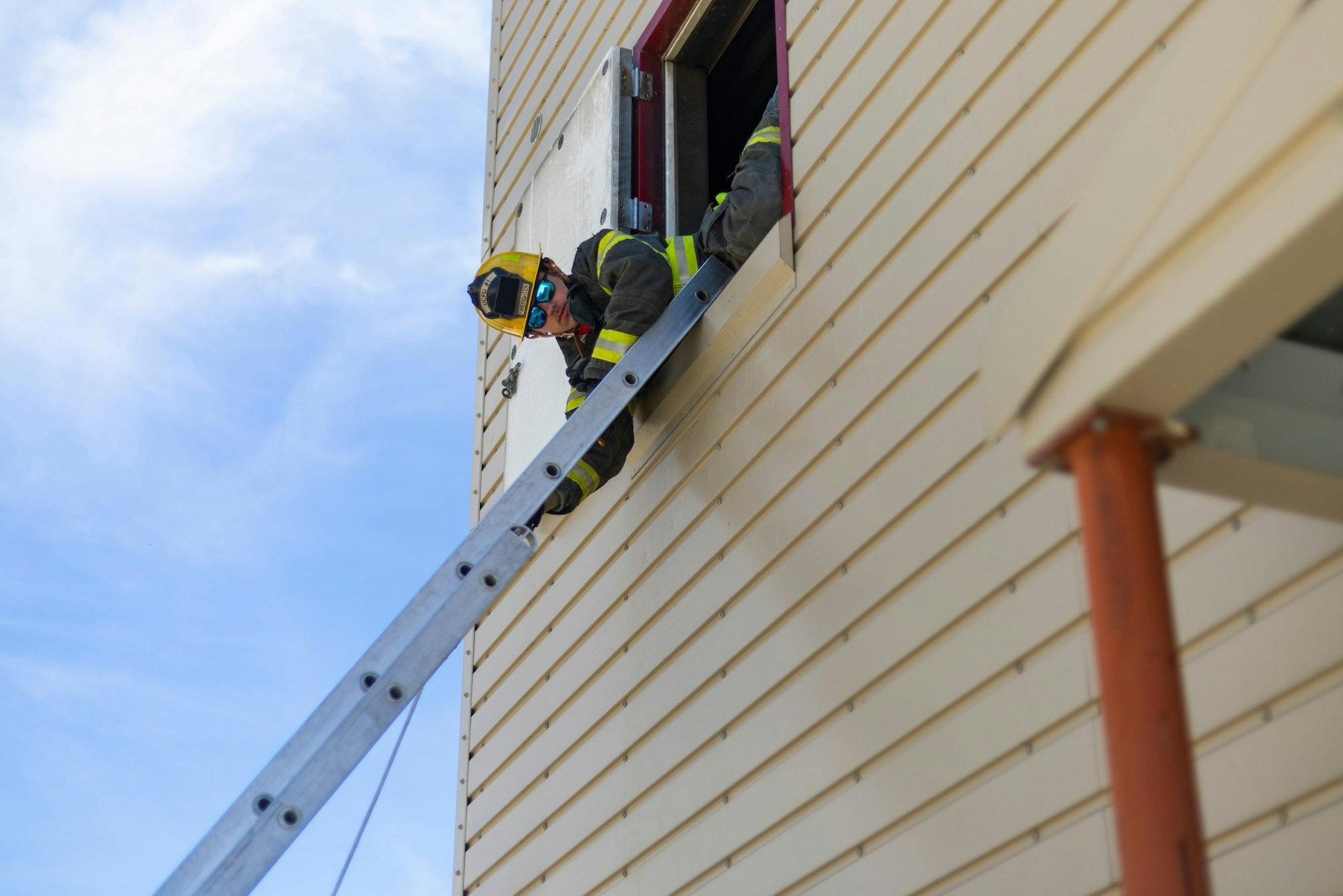 A firefighter in gear climbs a ladder through a building window.