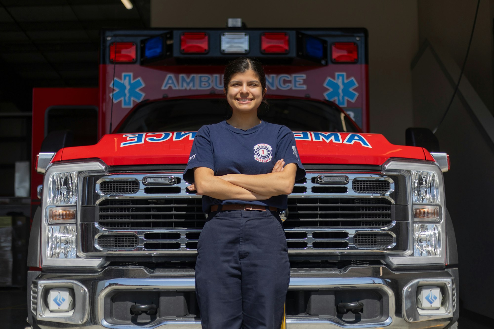 A smiling person stands confidently in front of a red ambulance with their arms crossed.