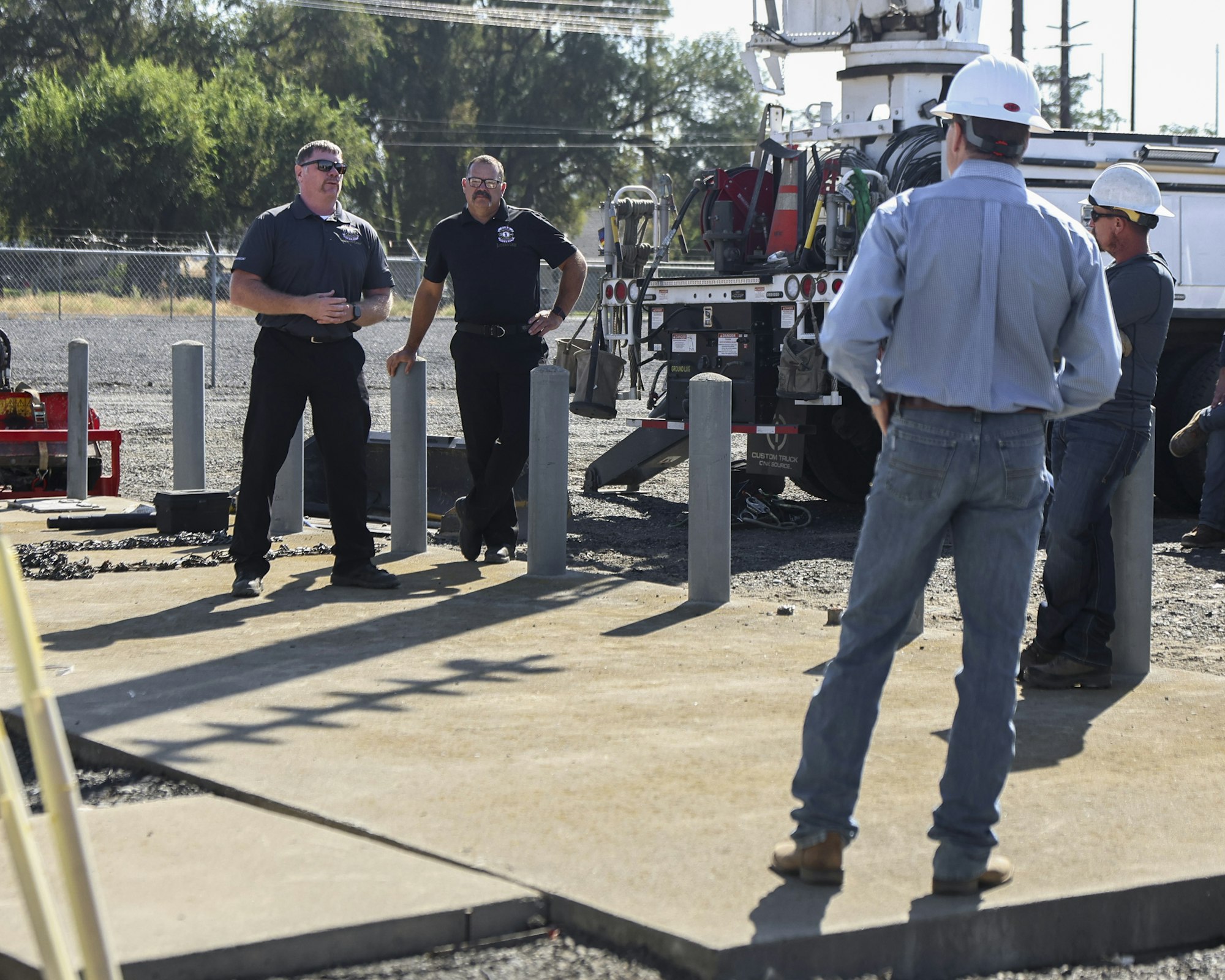 A group of workers in hard hats and uniforms talk next to utility equipment outdoors.