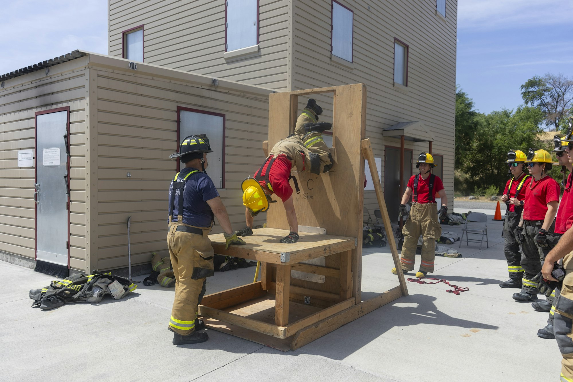 Firefighters training with an obstacle course; one climbs through a small window as others watch for guidance.