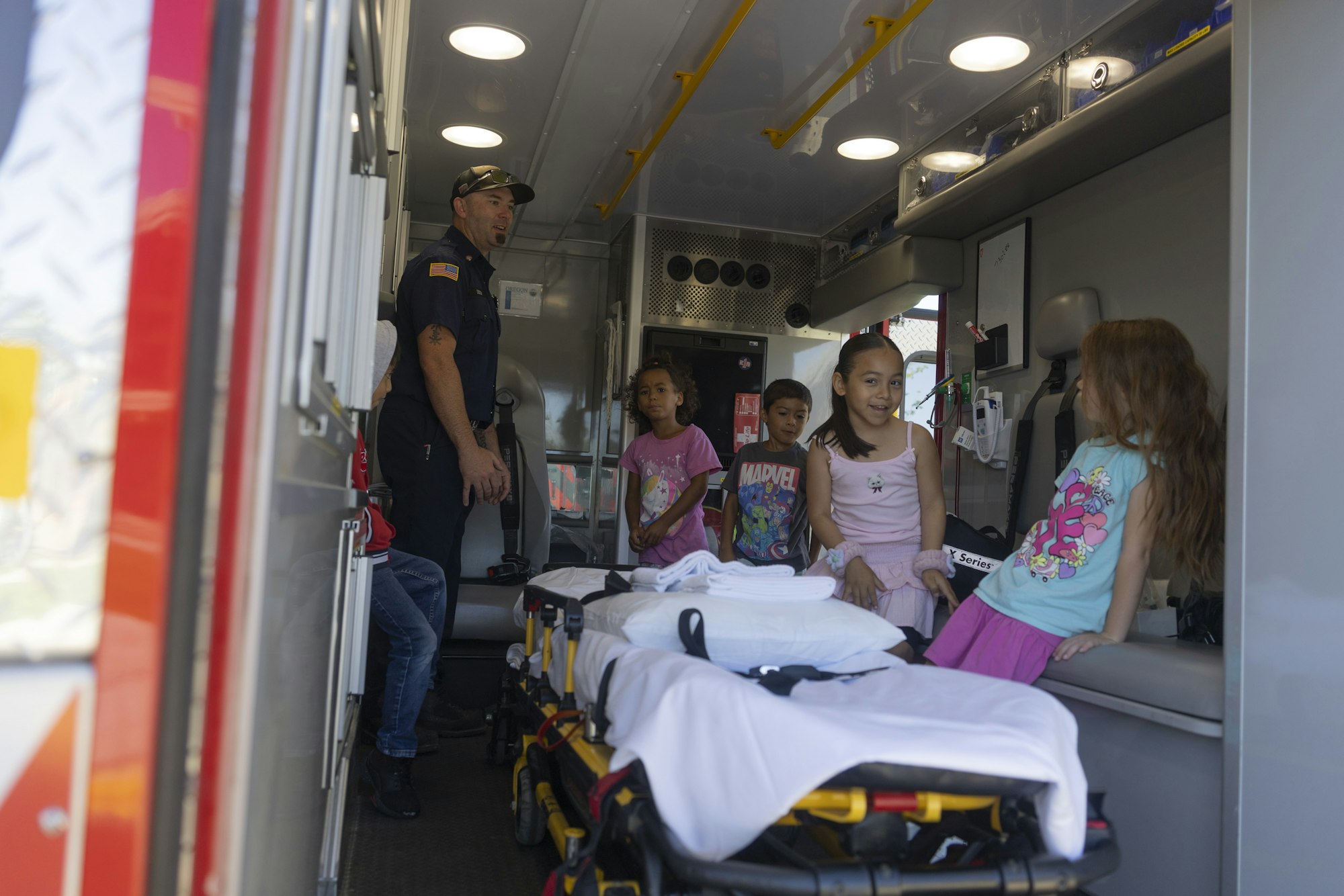 A paramedic shows an ambulance interior to a group of children gathered inside, with medical equipment visible.