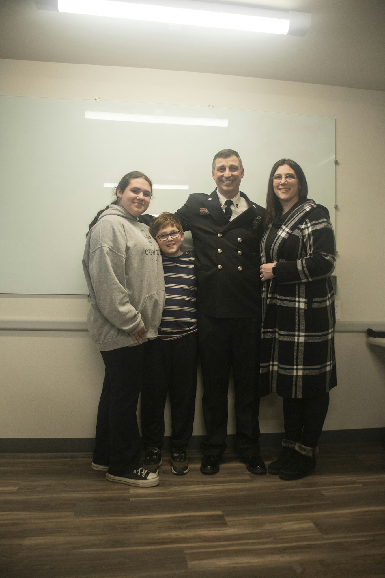 A group of four people smiling, includes one in a uniform and others in casual clothing, standing indoors.