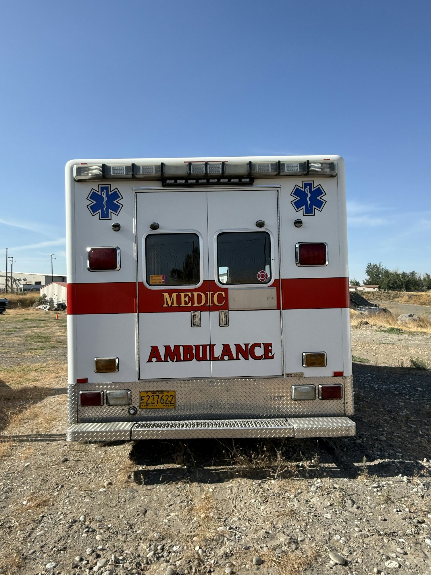 The rear of an ambulance with "MEDIC" and "AMBULANCE" text, parked on a gravel surface under a clear sky.