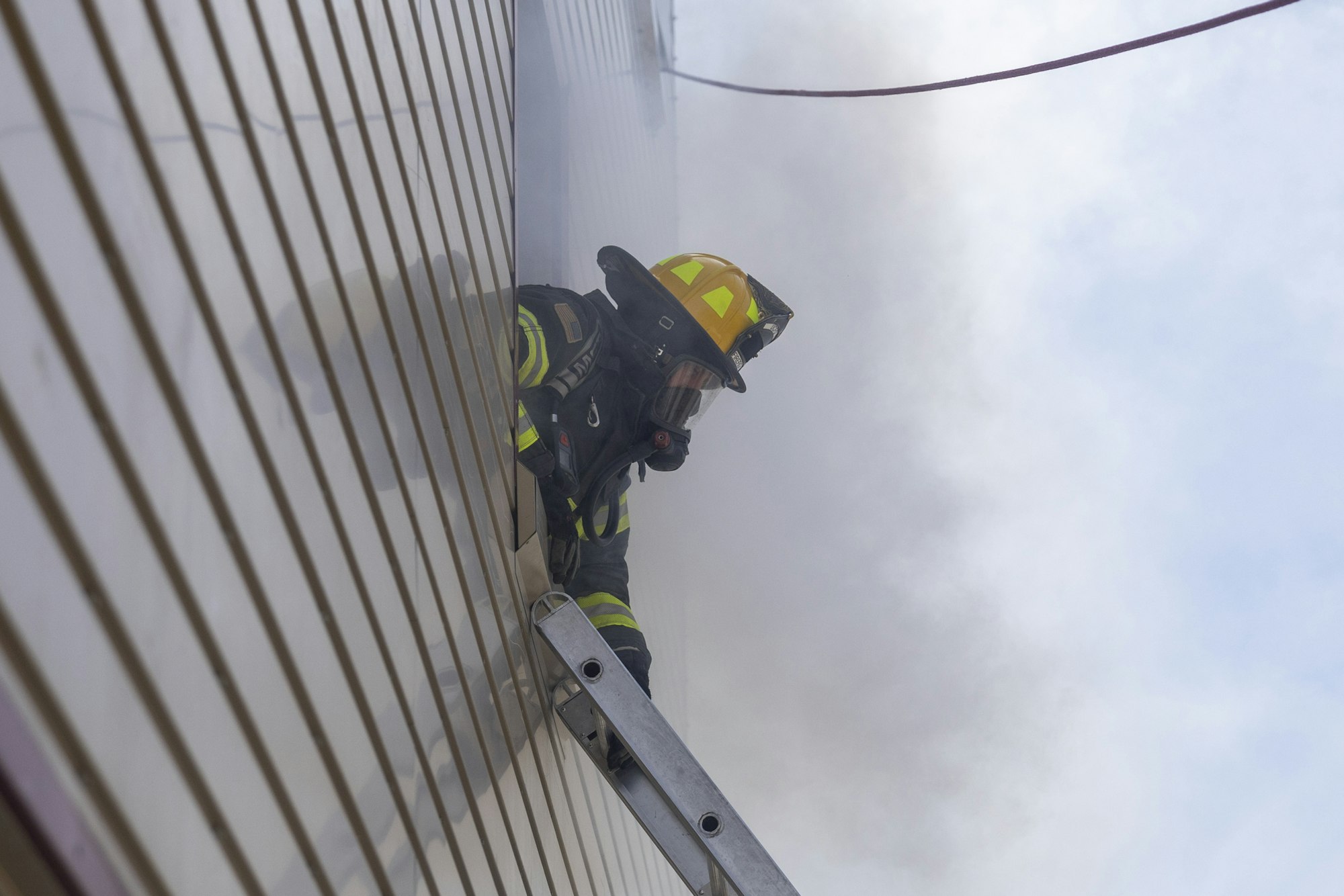 Firefighter on ladder at smoky building, wearing gear.