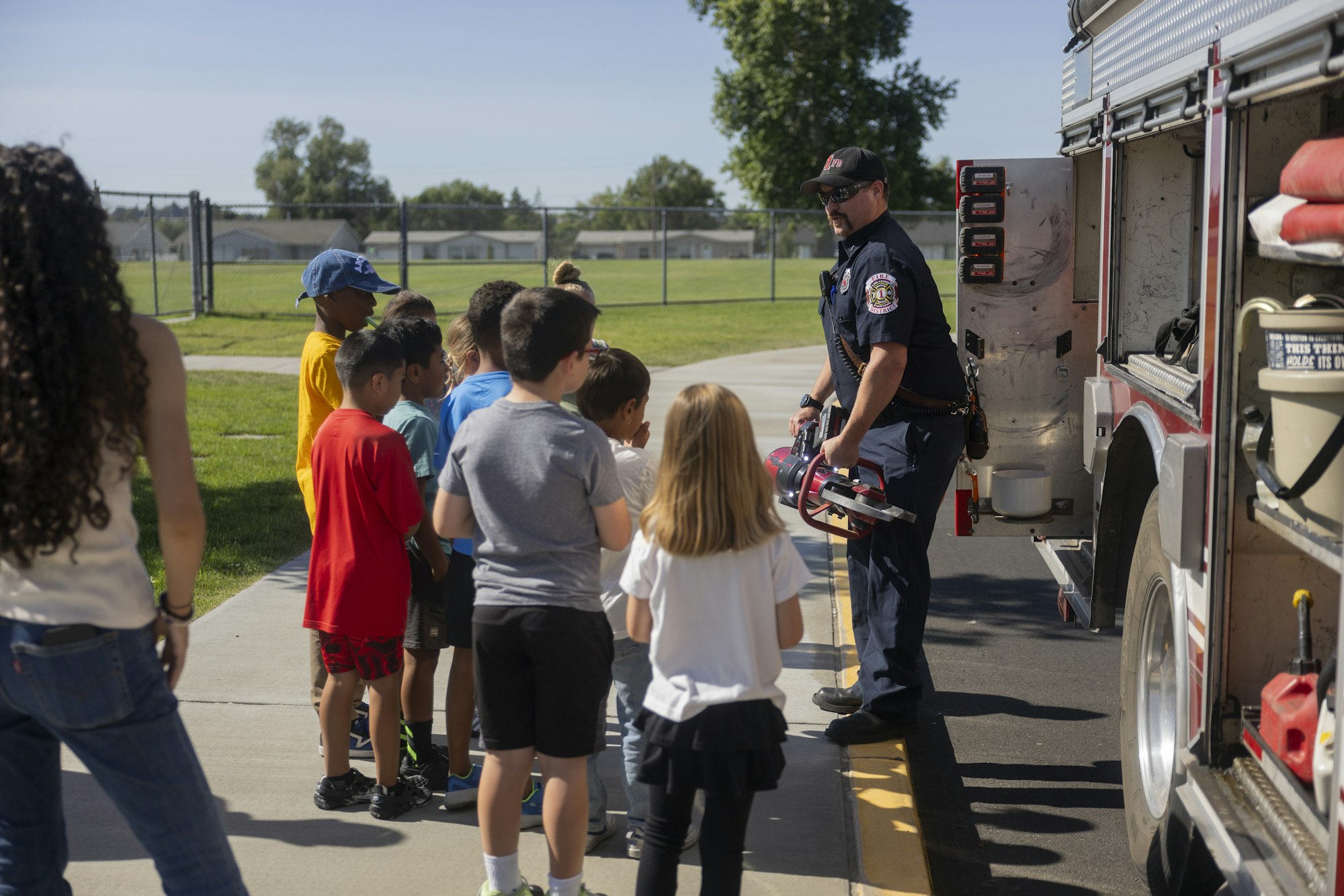 A firefighter showing tools to a group of children by a fire truck.