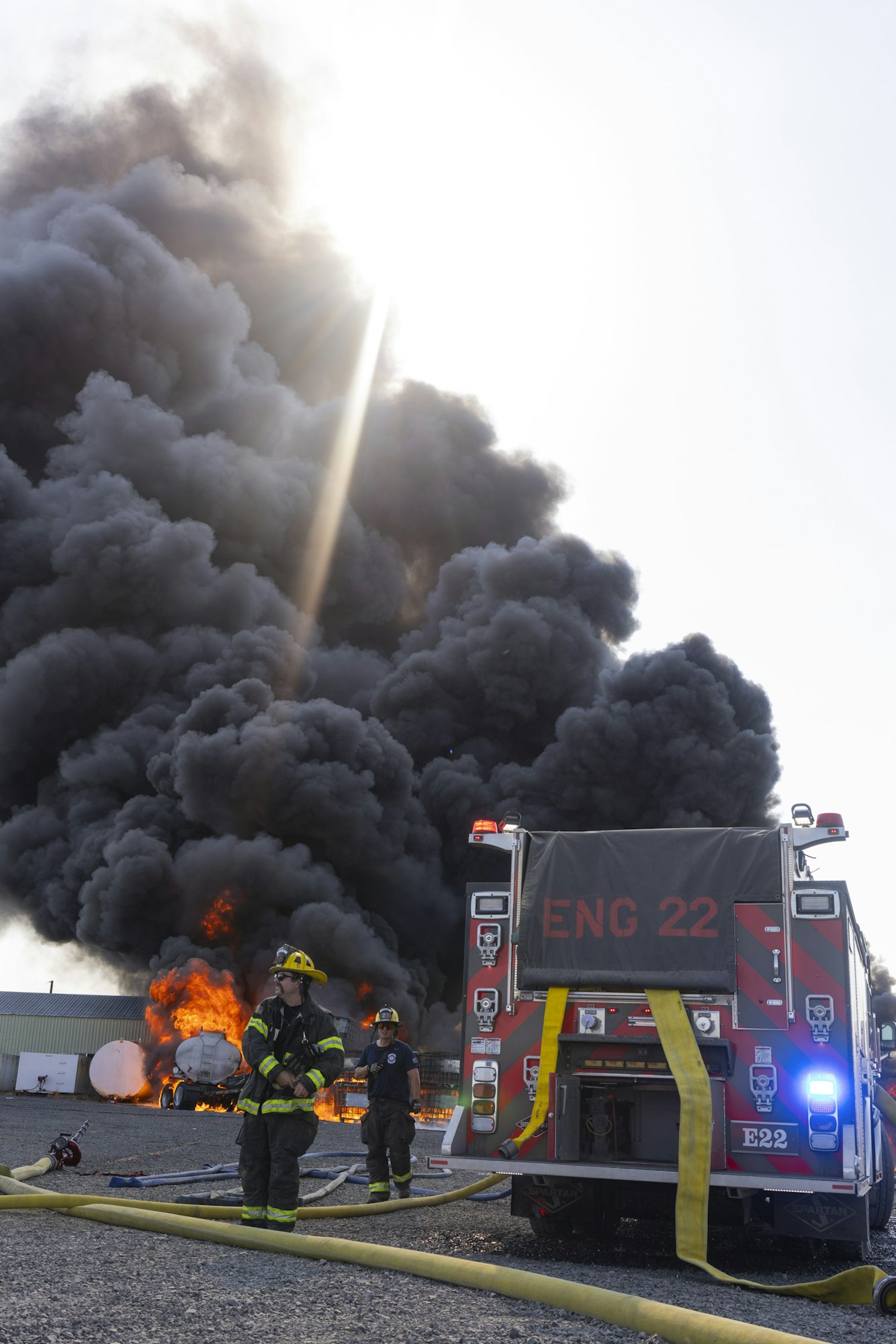 Firefighters and a fire truck at a large industrial blaze with thick smoke.