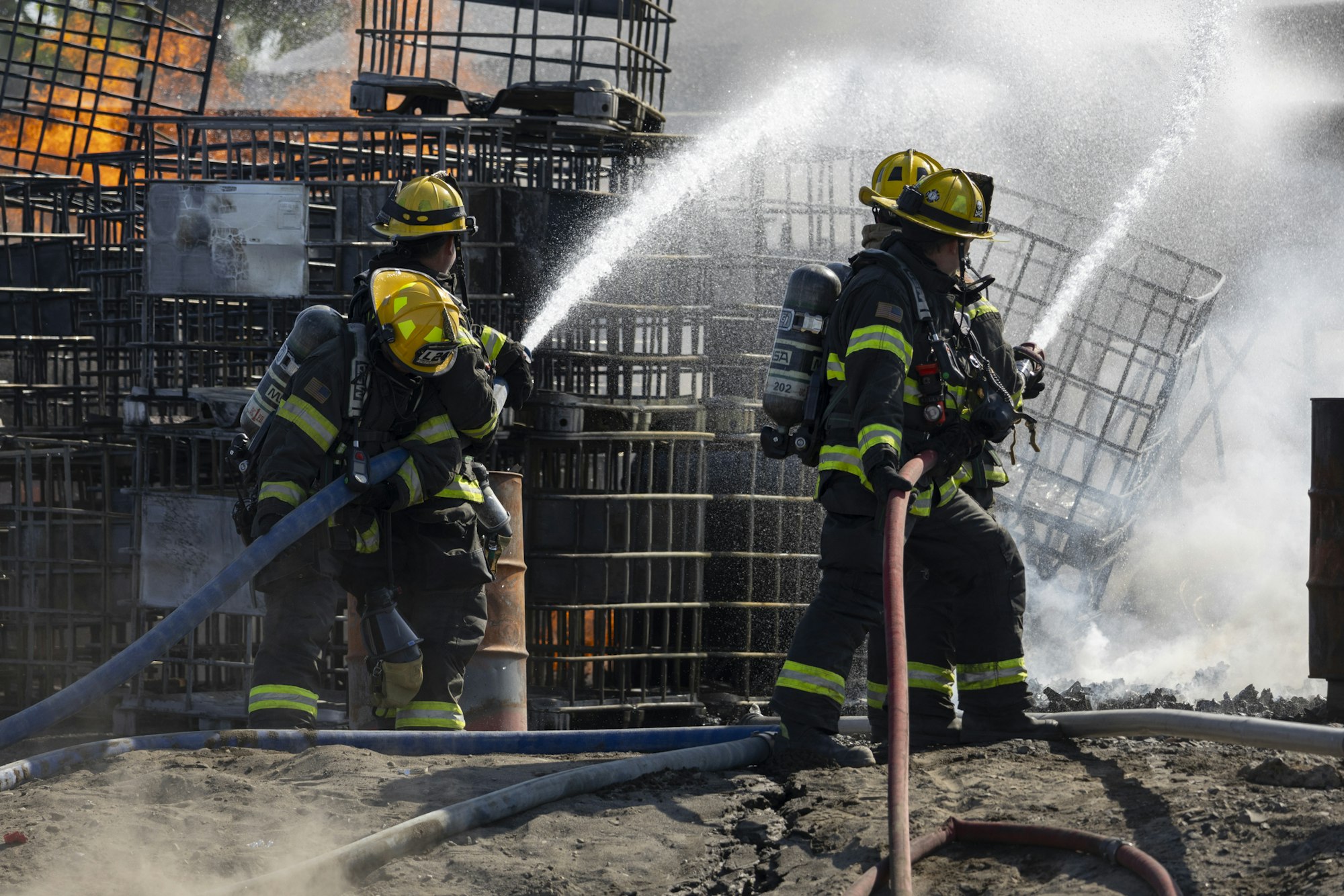 Firefighters in gear extinguishing a fire with hoses, smoke and flames visible.
