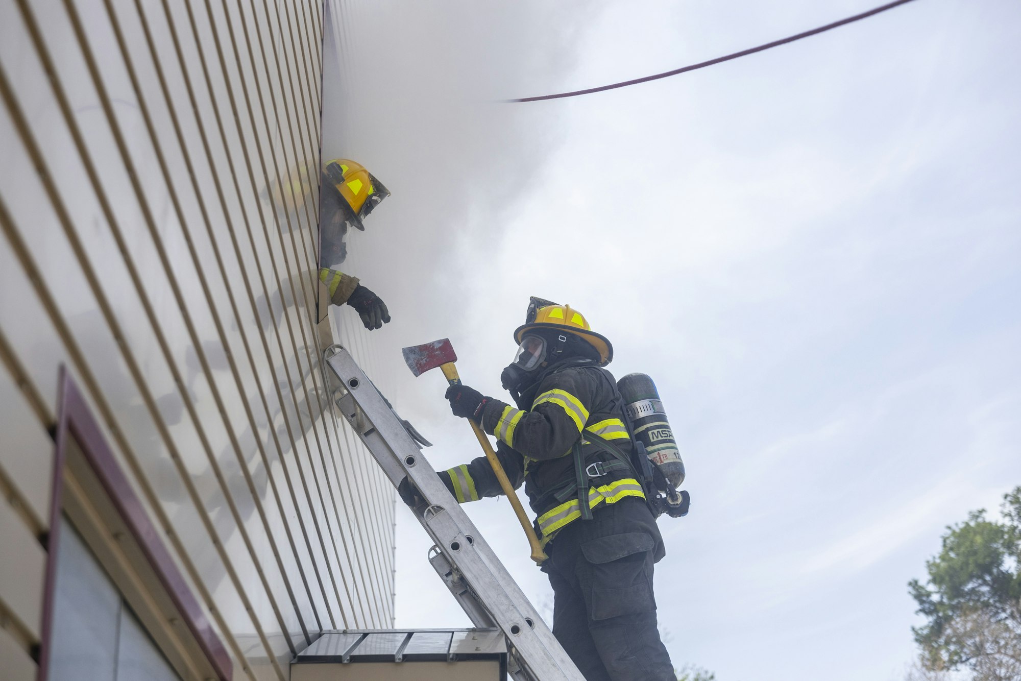 Two firefighters in gear, one on a ladder with an axe, working near a smoke-filled building.