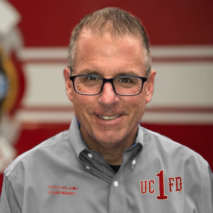 A person wearing glasses and a gray shirt with "UC 1 FD" text, standing in front of a blurred red and white background.