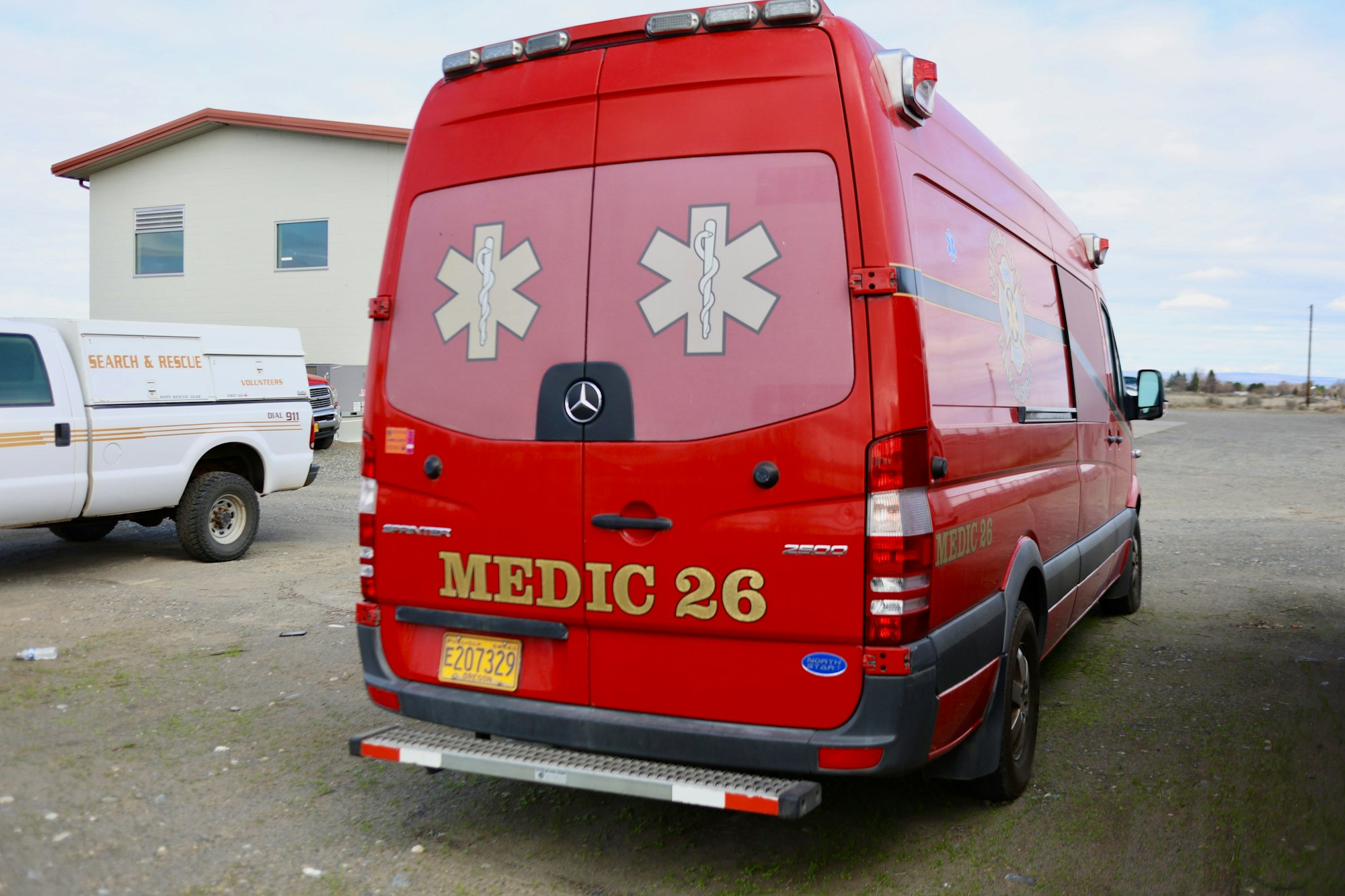 Red ambulance labeled "Medic 26" with medical symbols, parked alongside a search and rescue vehicle.