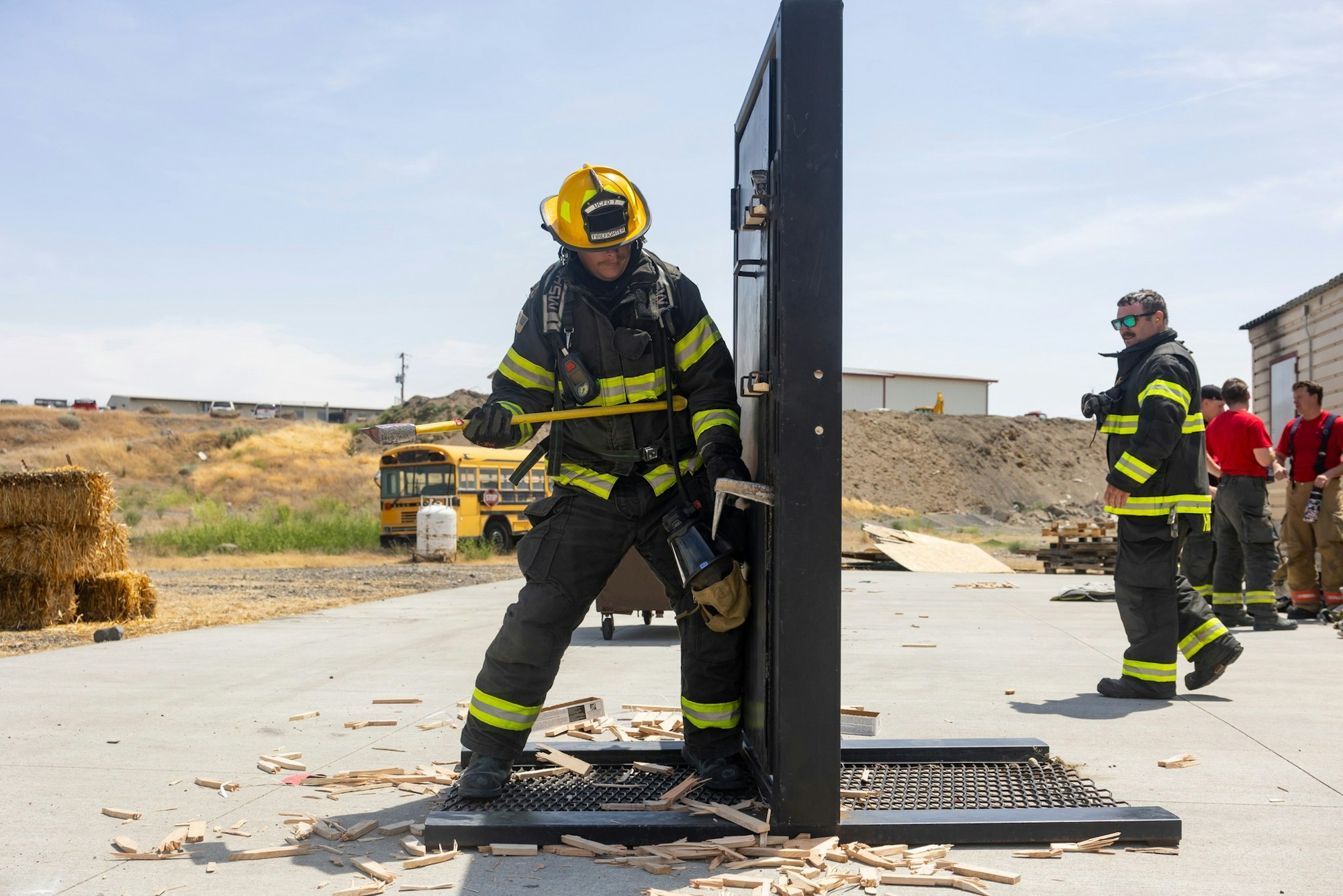 Firefighter breaking a door with an axe during training; others observe, wearing safety gear.