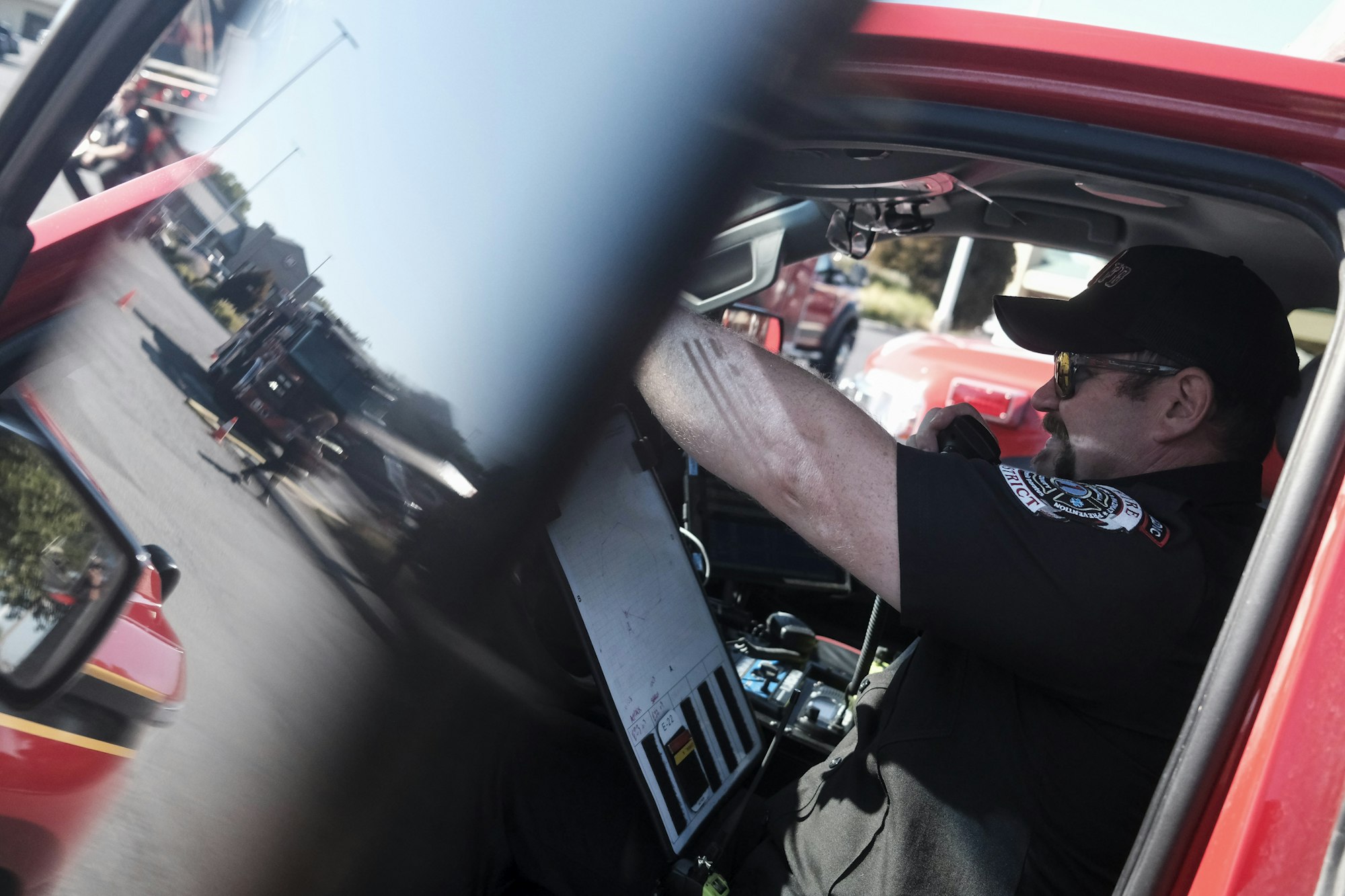A person in a vehicle, using a radio, wearing a uniform with insignia, and there's a reflection of a fire truck outside.