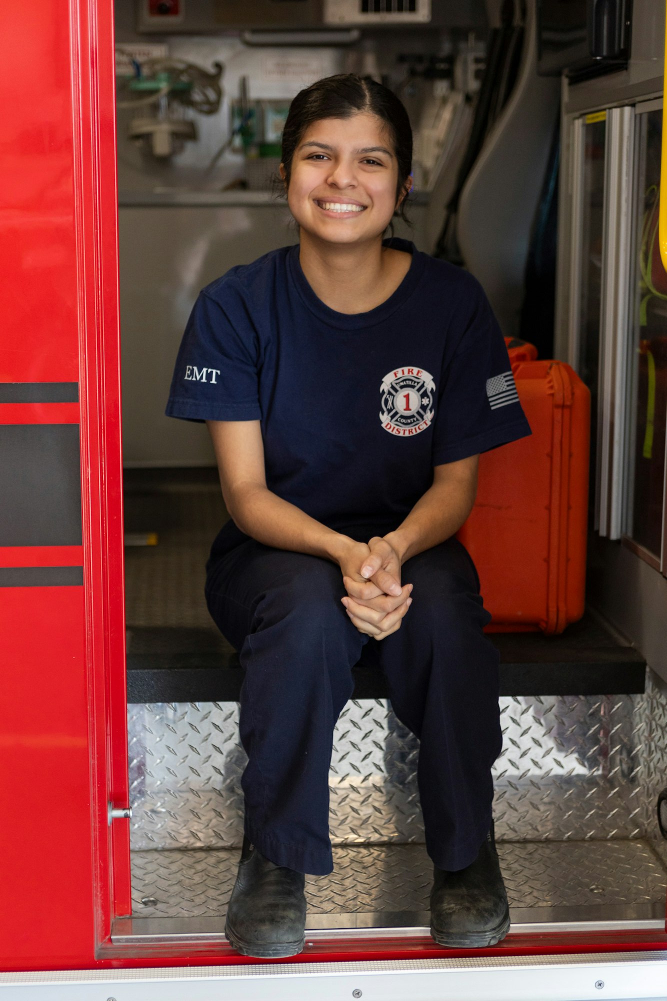 An EMT in uniform is sitting inside an ambulance, smiling.