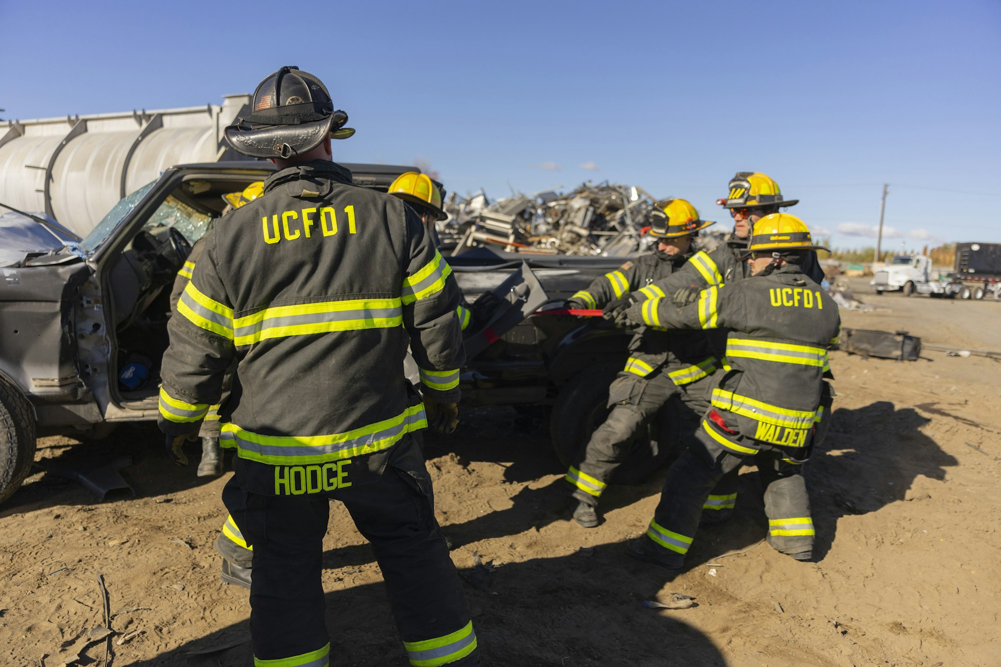 Firefighters in gear work on a damaged vehicle in a scrapyard.
