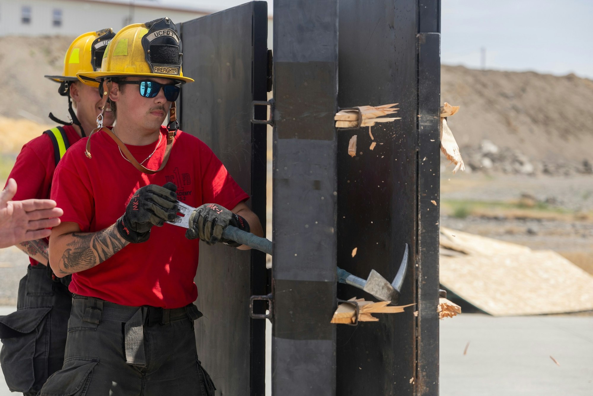 Firefighters in training, wearing helmets and red shirts, use tools to break through a wooden barrier outdoors.
