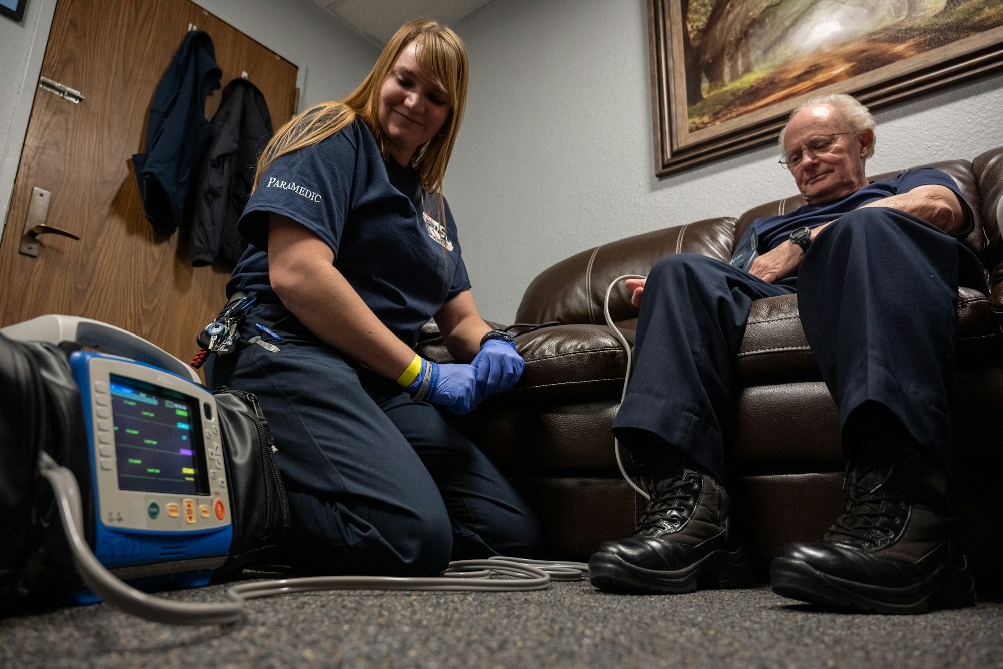 A paramedic kneels next to an older man on a couch, using medical equipment.