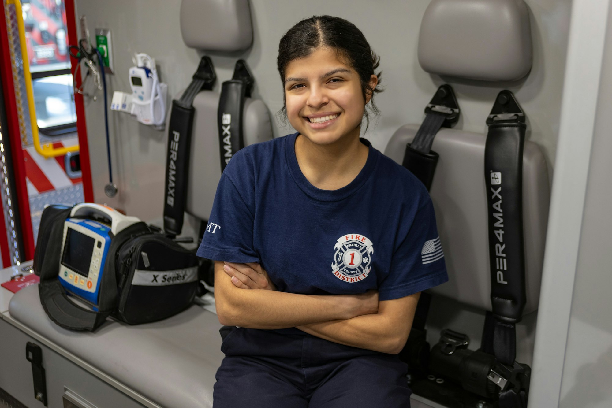 A person in a fire department shirt sitting in a vehicle with medical equipment nearby, smiling at the camera.