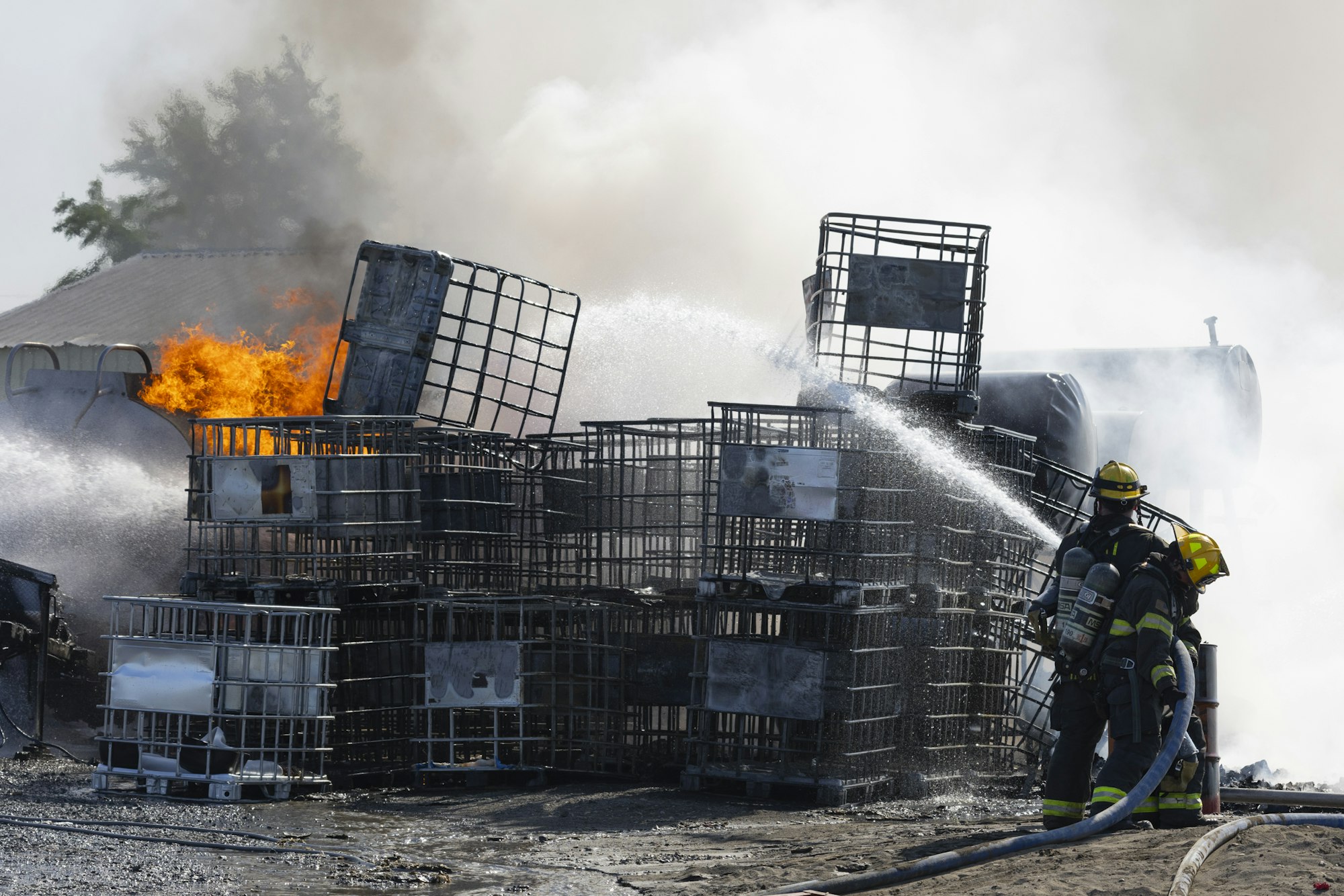 Firefighters extinguishing a fire in industrial containers with hoses spraying water.