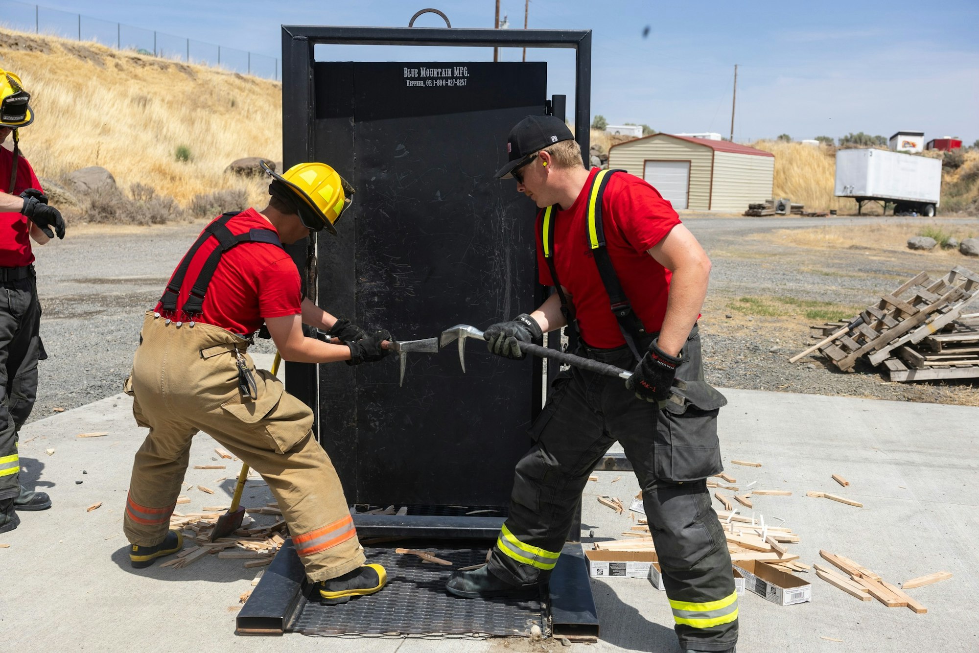 Two firefighters practice forcible entry on a training door outside, using tools and wearing safety gear.