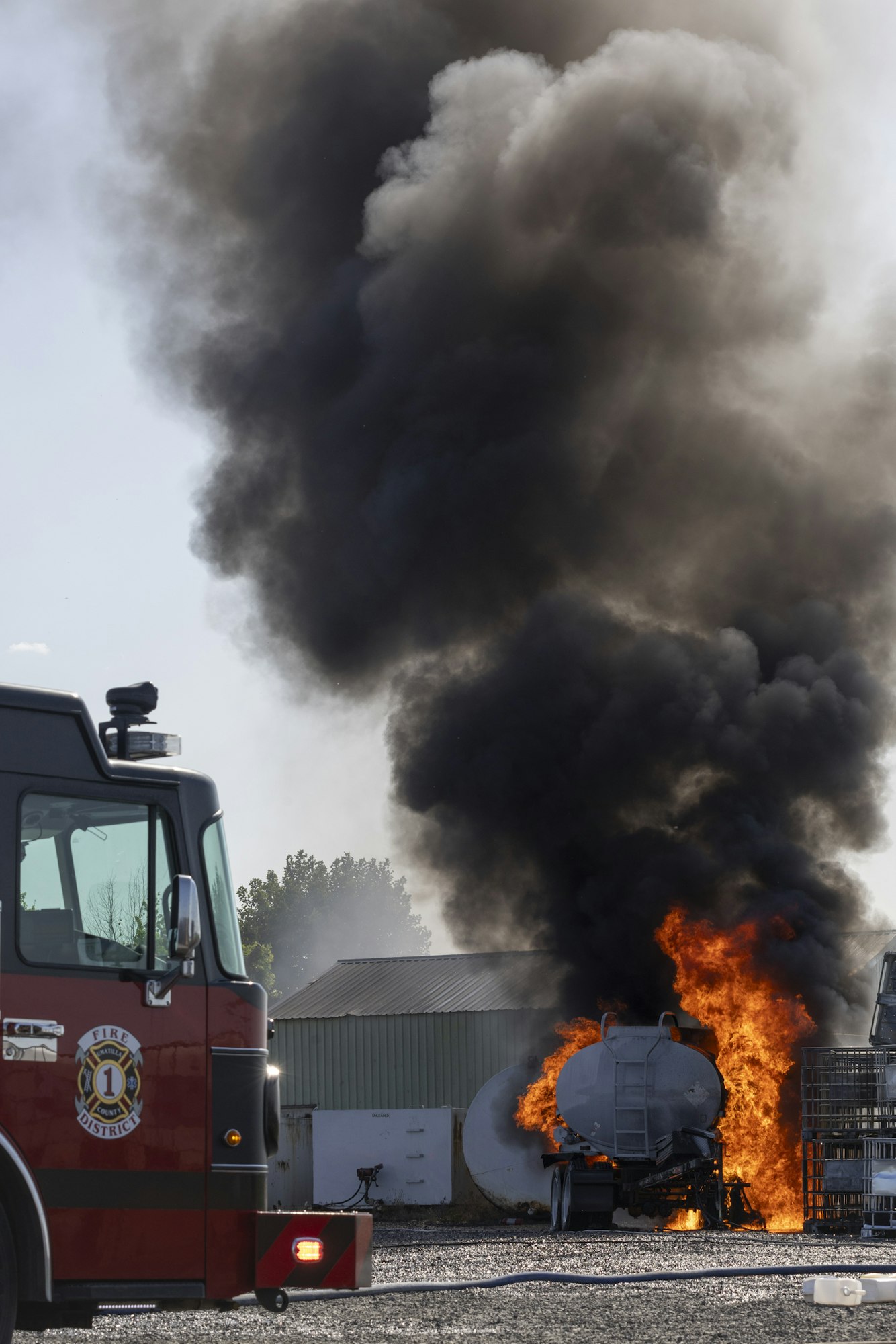 Fire truck near a large fire with black smoke and flames engulfing a tanker truck or similar vehicle.