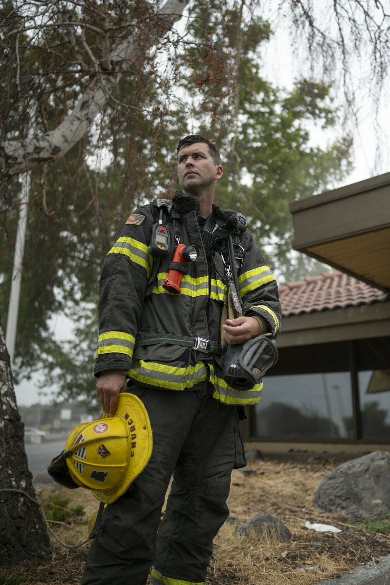 A firefighter in gear holds a yellow helmet, standing outdoors near trees and a building.