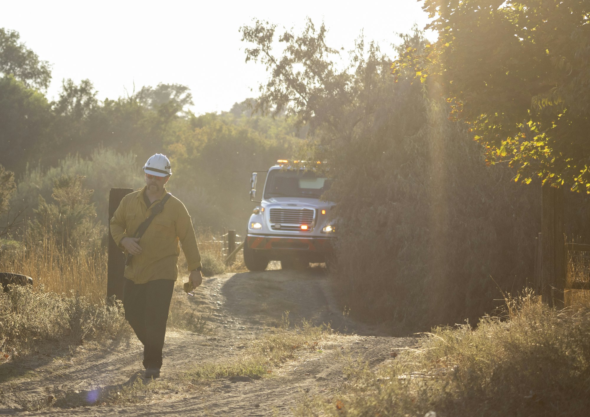 A person in safety gear walks on a dirt path with a truck in the background, surrounded by foliage.