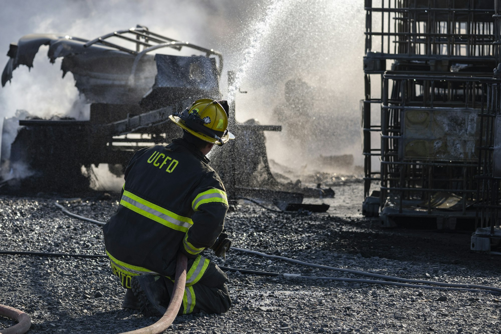 A firefighter spraying water on smoking debris and wreckage, wearing gear labeled "UCFD."