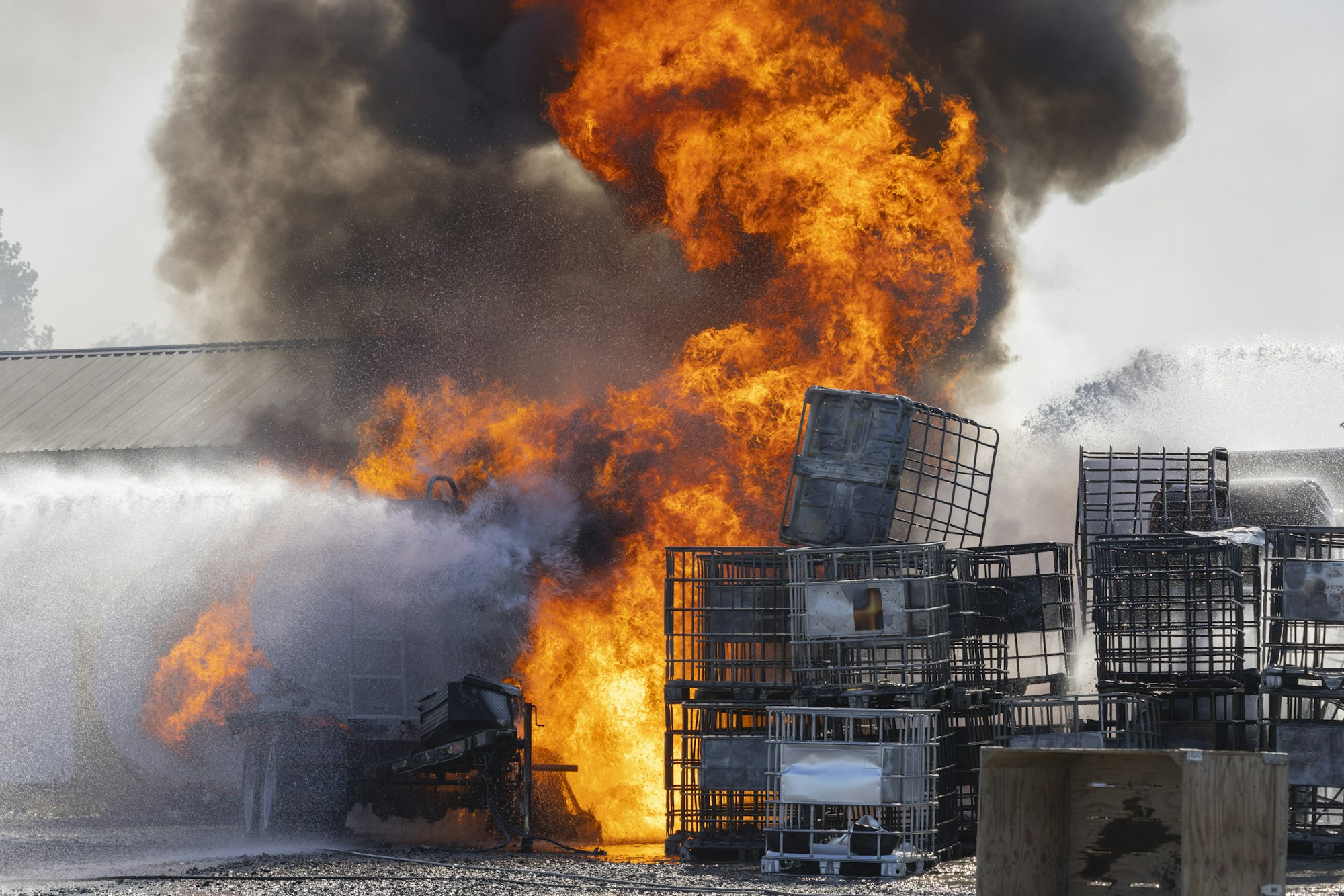 A massive industrial fire with intense flames and black smoke, surrounded by several metal containers and splashes of water.