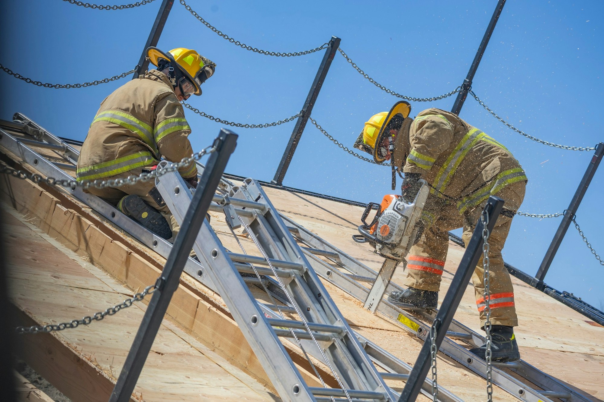 Two firefighters in gear work on a sloped roof with ladders and chainsaws under a clear blue sky.