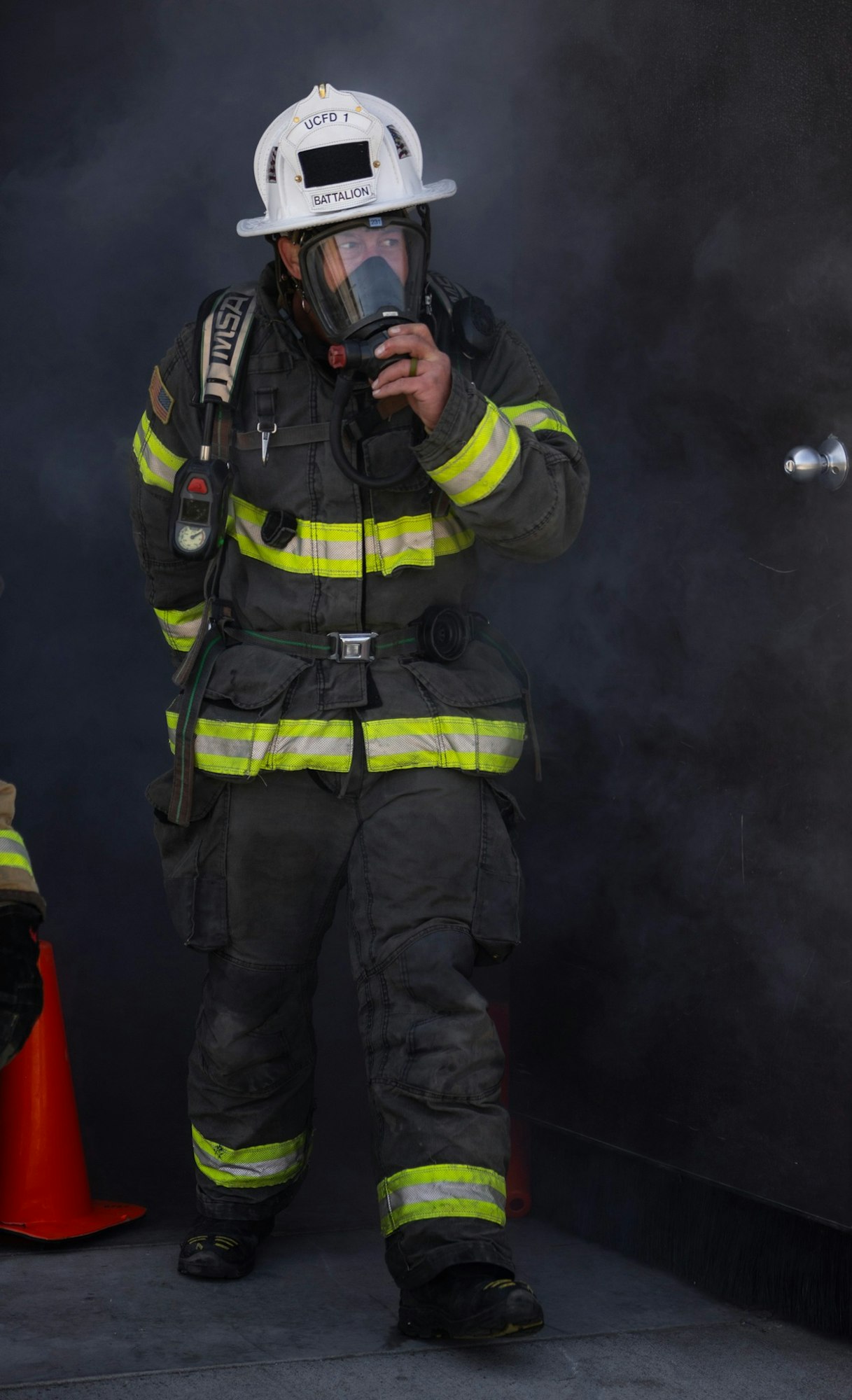 A firefighter in full gear and helmet, holding a mask, walking through smoke with a reflective stripe uniform.