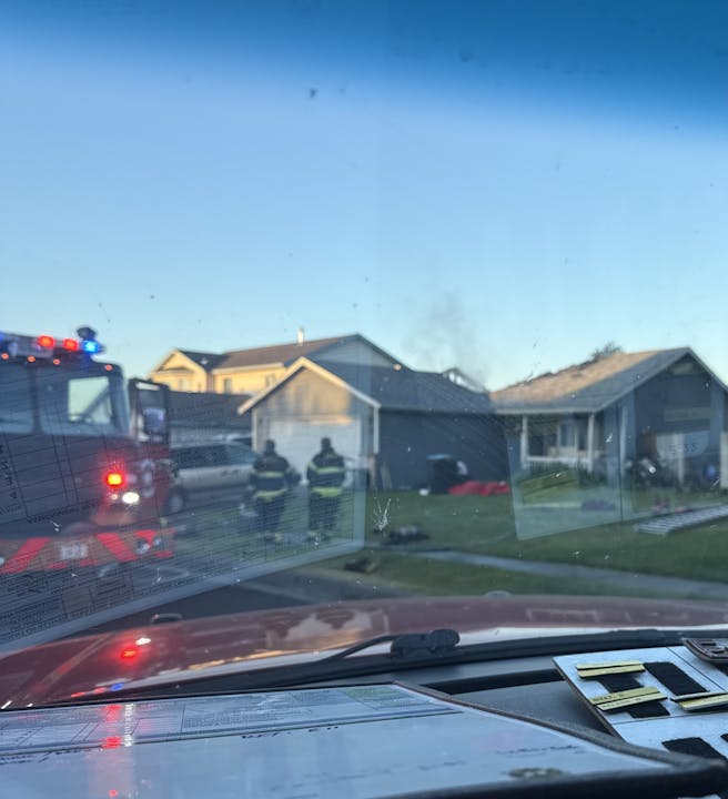 Firefighters and a fire truck outside a house, viewed through a car windshield.