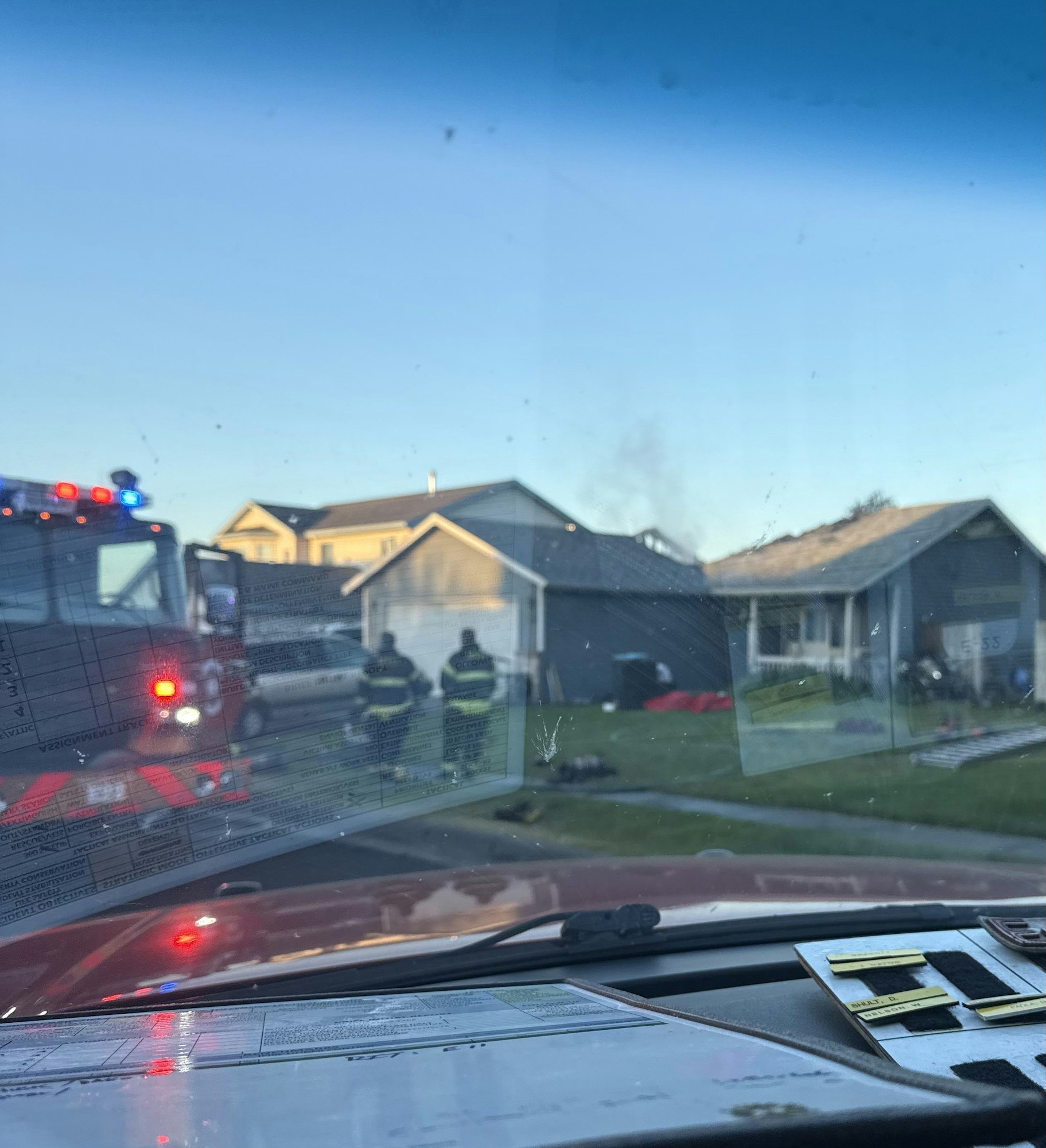 Firefighters and a fire truck outside a house, viewed through a car windshield.