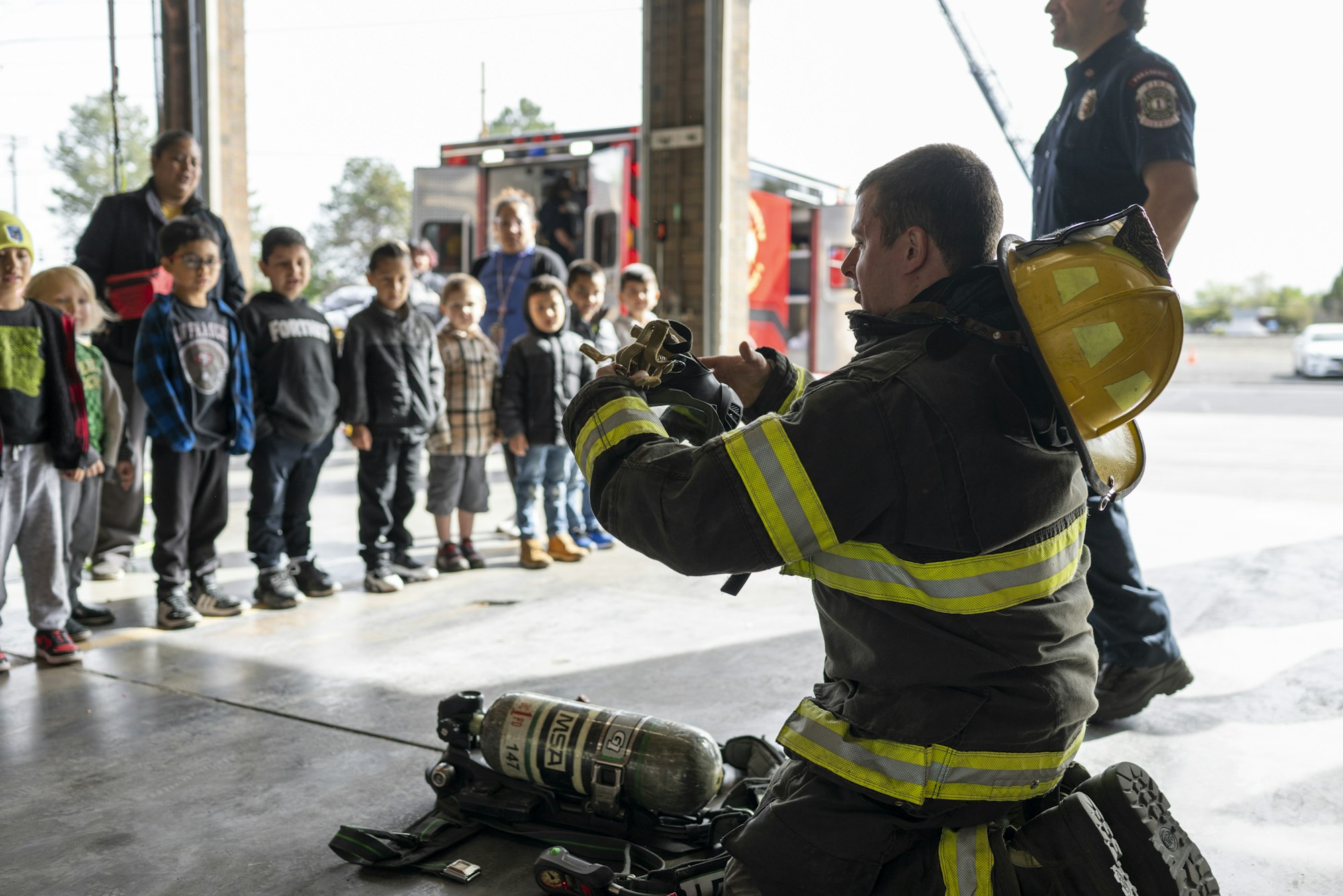 Firefighter demonstrating equipment to a group of children in a fire station.