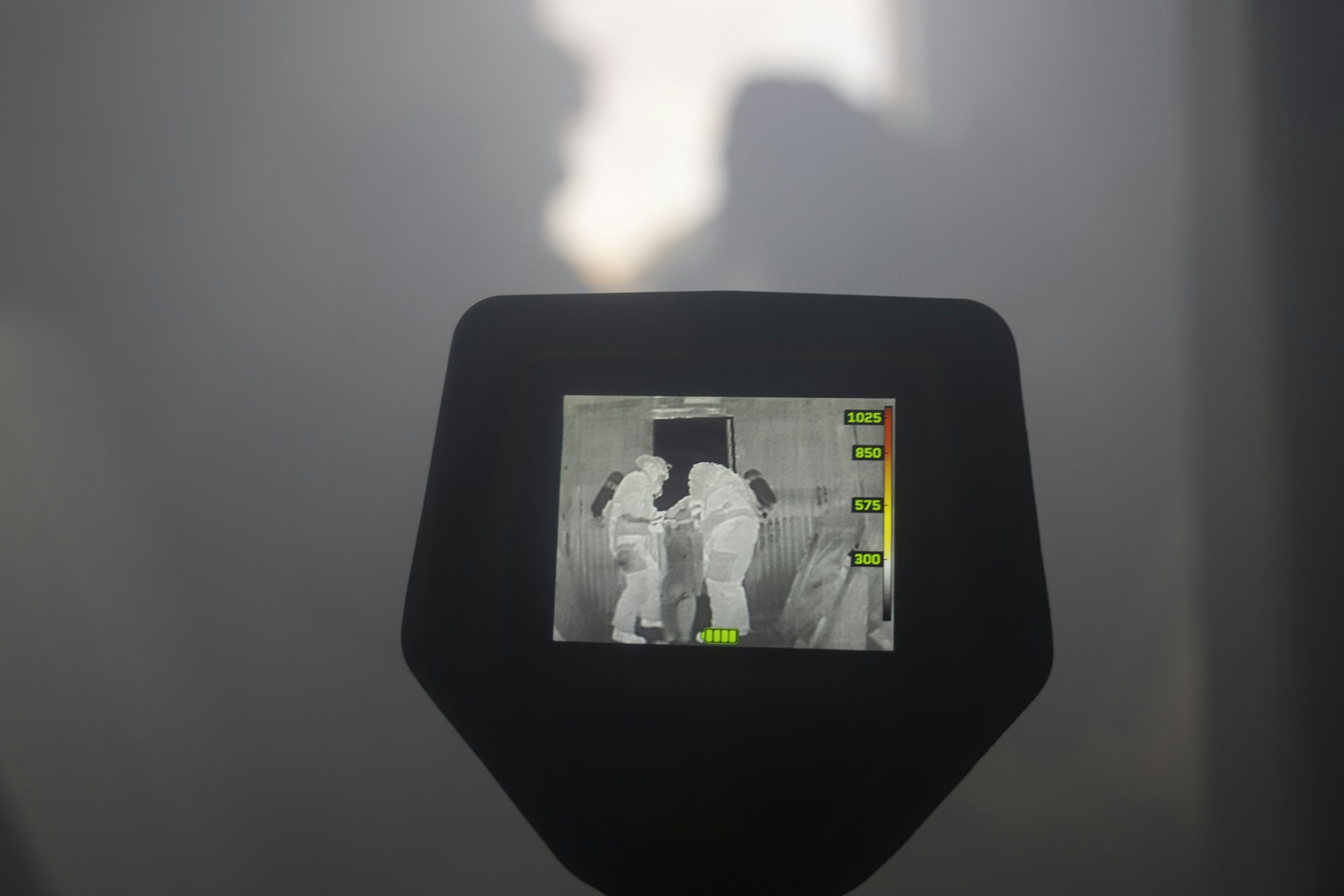 A thermal imaging device shows two figures in a smoky or foggy environment.