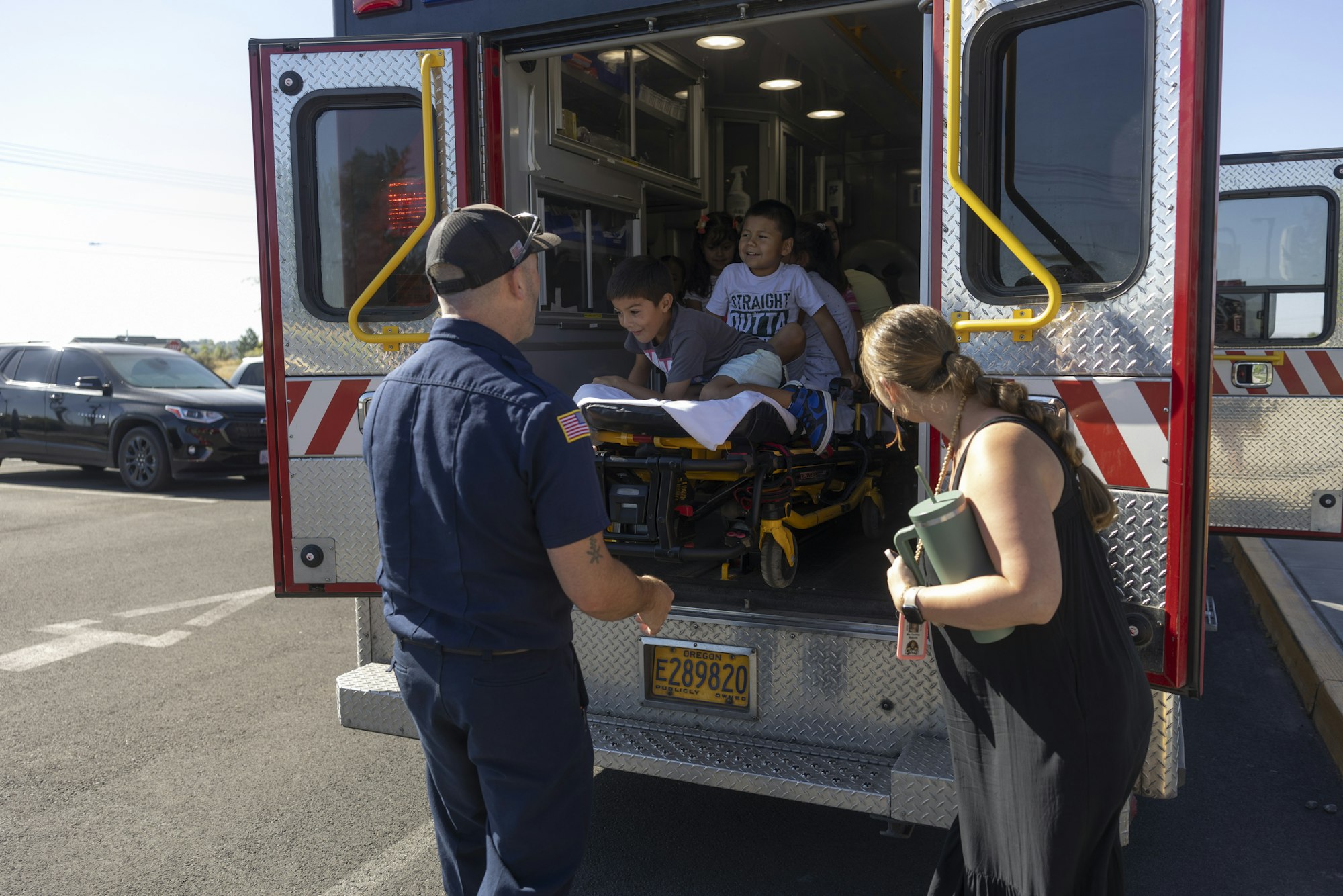A firefighter stands by an ambulance where children are playing inside, while a woman watches from outside.