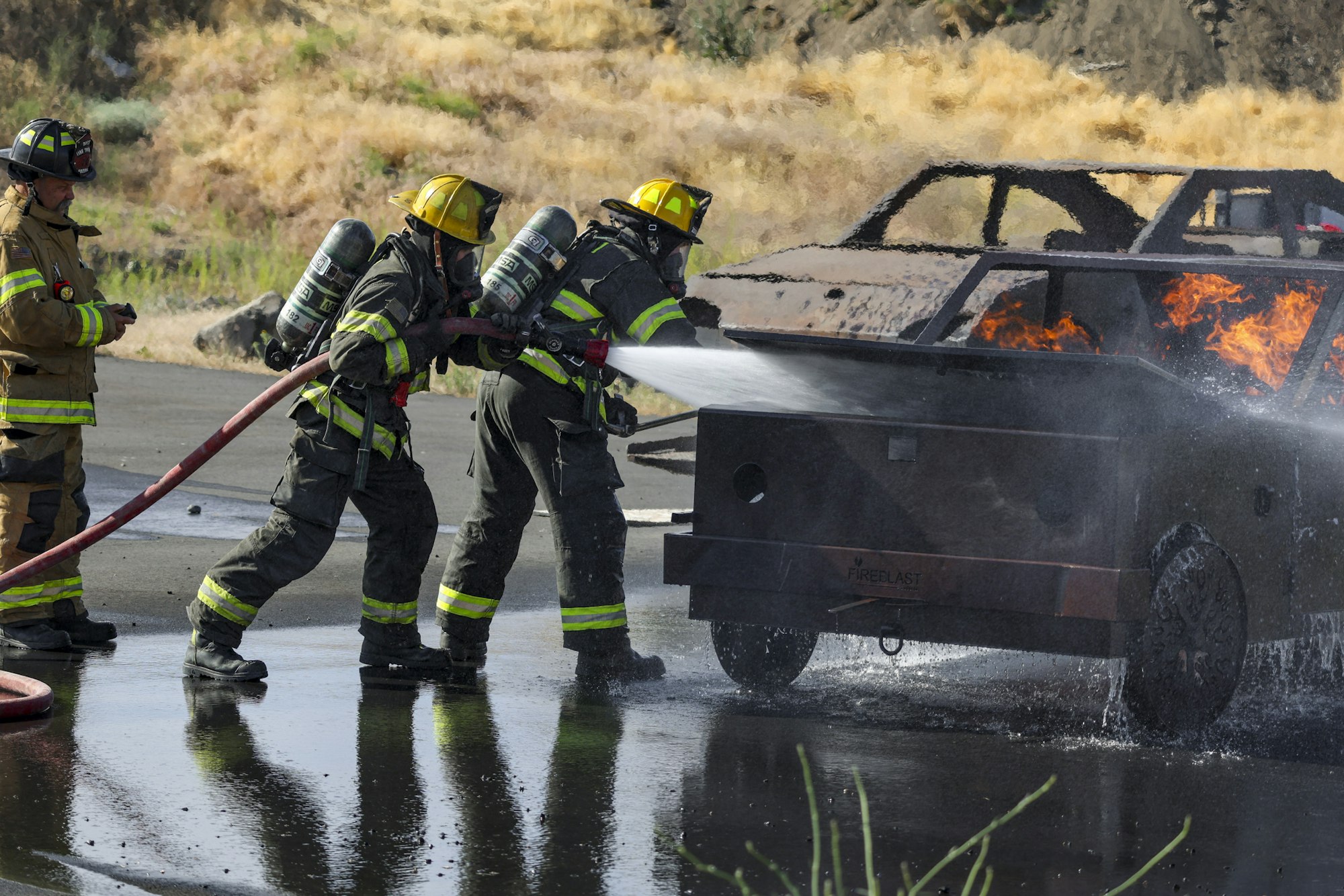 Firefighters extinguishing a car fire using a hose, wearing full gear and helmets.
