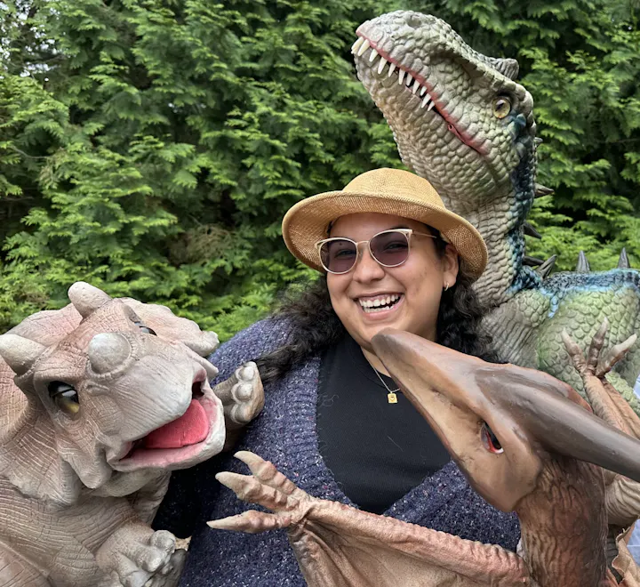 A person is joyfully posing with several dinosaur toys amidst green foliage.