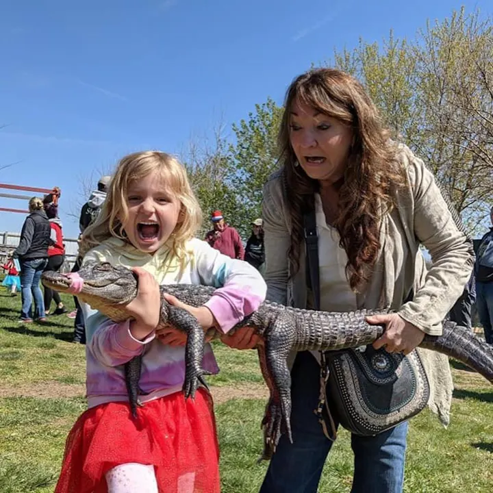 A girl excitedly holds a small alligator while a surprised woman assists her, surrounded by people outdoors.