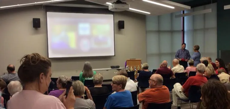 A group of people in a room with a blurred projection on a screen, suggesting a lecture or meeting setting.