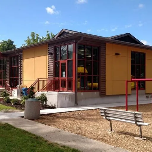 A modern building with large windows, yellow and brown walls, a bench, and a trash bin outside under a clear sky.