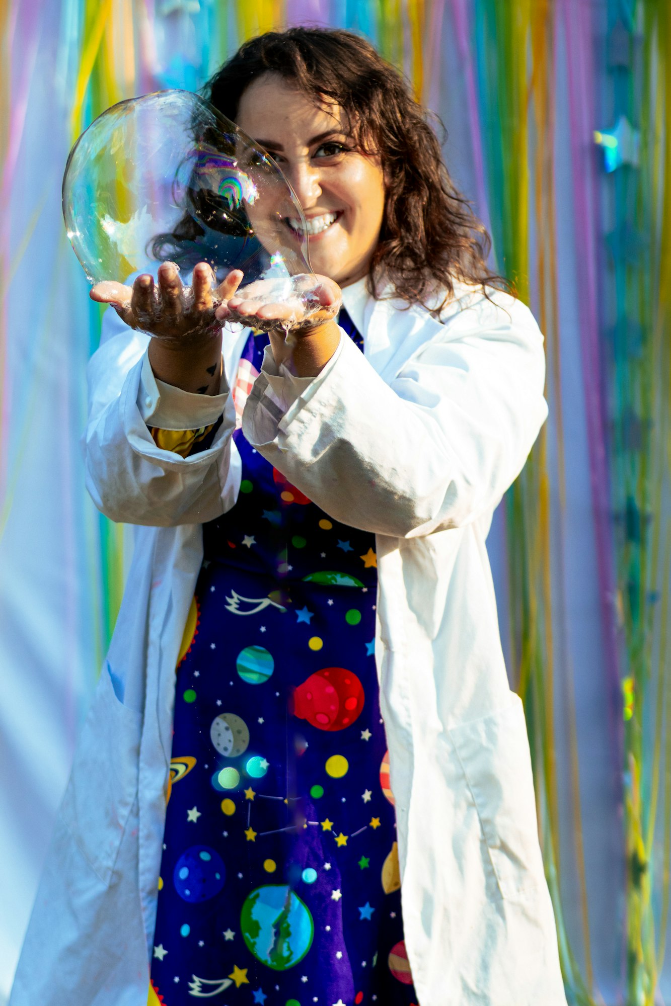 A person in a lab coat holding a large, iridescent bubble, with a colorful backdrop of streamers.
