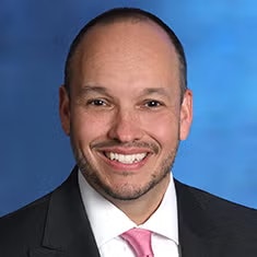 Smiling man in a suit and pink tie with a blue background.