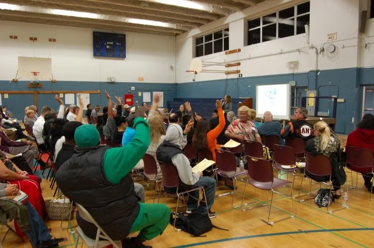 A group of people in a gymnasium raising their hands during a presentation or meeting.