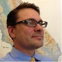 A person wearing glasses and a blue shirt with a tie, looking to the side, in front of a map.