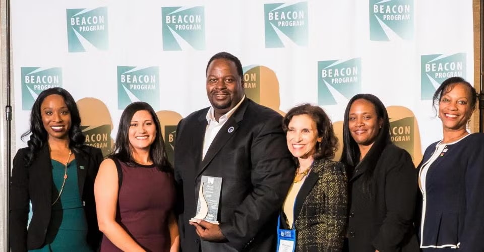 Seven people posing with an award in front of a "Beacon Program" banner.