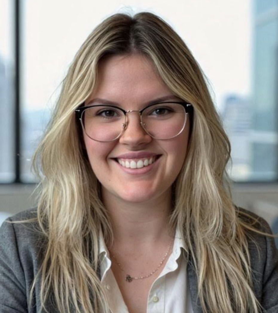 A person with long blonde hair, glasses, and a gray blazer smiles indoors.