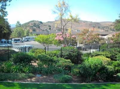 Lush garden with trees, a metal fence, and a view of a parking lot and hills in the background.