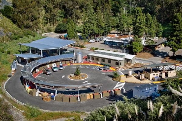 Aerial view of a circular recycling center with parked vehicles, a central plant island, and several structures surrounded by trees.