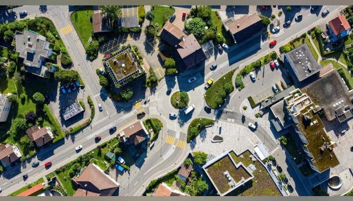 Aerial view of a roundabout with cars, surrounded by buildings, greenery, and parking areas.